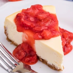 Slice of New York Style Cheesecake on a white plate with strawberry sauce on top.