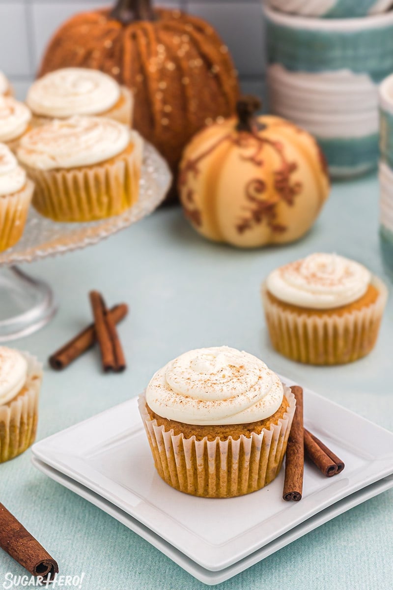 Portrait style photo of pumpkin spice cupcake on white square plate with other cupcakes and fall decor in the background.