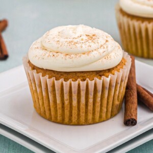 Pumpkin spice cupcake sitting on two stacked square white plates with two cinnamon sticks to the right of it.