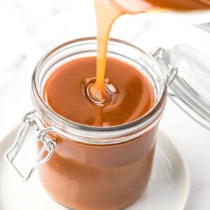 Pouring caramel sauce into a glass jar that is placed on a white plate.