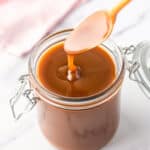 A spoon covered in and dripping caramel sauce over a glass jar of caramel sauce.