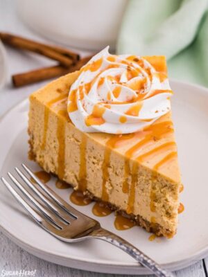 Slice of pumpkin spice cheesecake topped with whipped cream and cinnamon on a plate with a fork beside it.