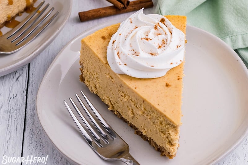 Slice of pumpkin spice cheesecake topped with a rosette of whipped cream and cinnamon set on a round white plate with a fork next to it.
