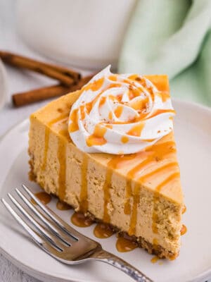 Slice of pumpkin spice cheesecake topped with whipped cream and cinnamon on a plate with a fork beside it.