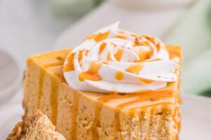 Slice of pumpkin spice cheesecake topped with whipped cream and caramel sauce with a bite set next to the slice.