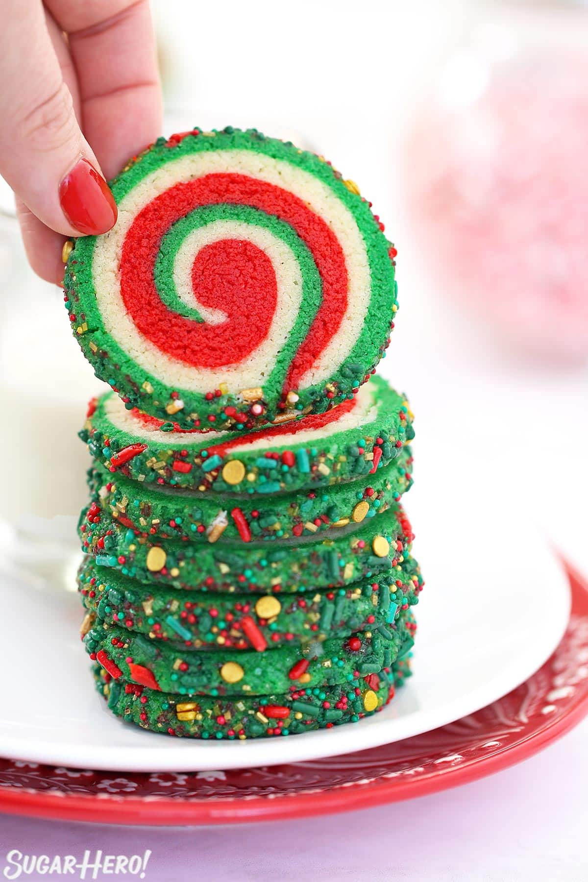 Hand with red fingernail lifting up a Christmas pinwheel cookie from a stack of cookies.