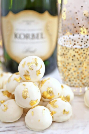 A dozen champagne white chocolate truffles in a pile with a glass of champagne and bottle of champagne in the background.