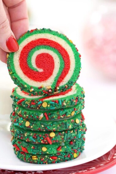 Hand with red fingernail lifting up a Christmas pinwheel cookie from a stack of cookies.