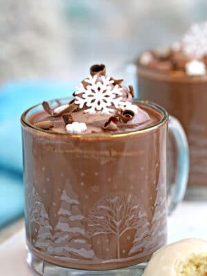Peppermint Hot Chocolate in a clear glass mug topped with cocoa whipped cream and edible snowflakes.