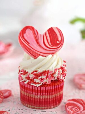SwA cupcake with white buttercream, pink and red sprinkles around the sides, and a pink and red swirled heart on top.