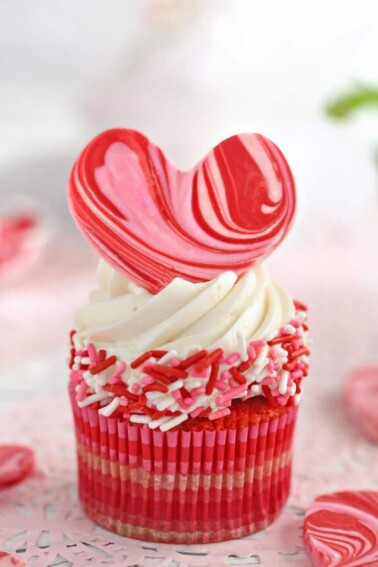 SwA cupcake with white buttercream, pink and red sprinkles around the sides, and a pink and red swirled heart on top.