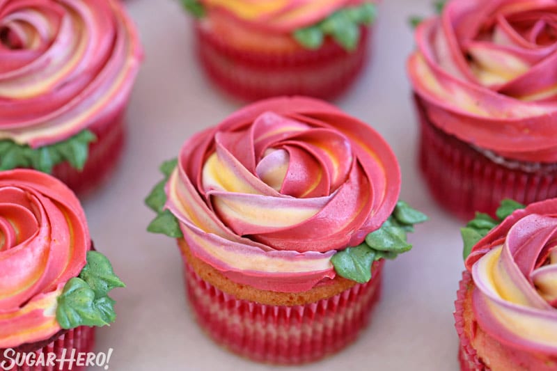 Group of pink and yellow rosette cupcakes with green leaves around the edges.