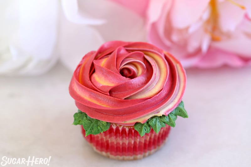 Single pink and yellow rosette cupcake with leaves around the edges, with pink and white flowers in the background.