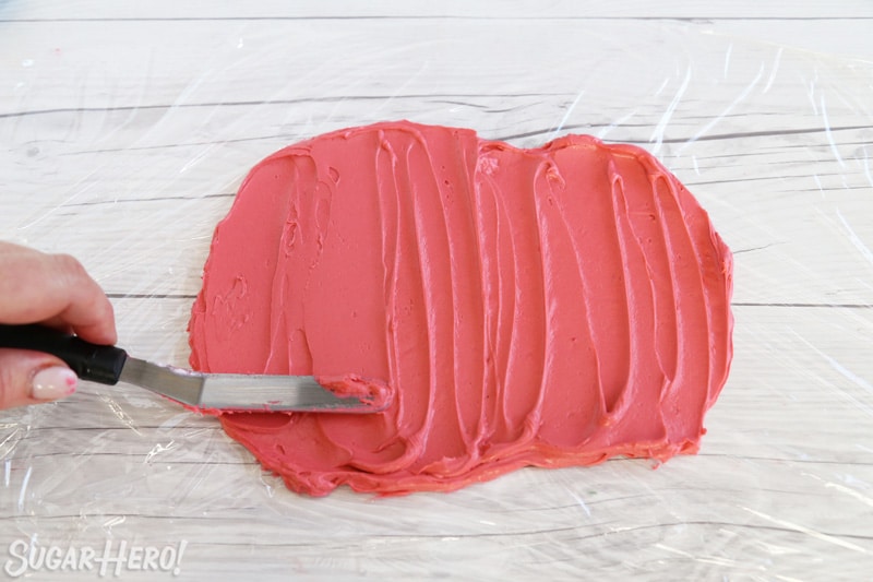 A small spatula spreading dark pink buttercream on a layer of plastic wrap.