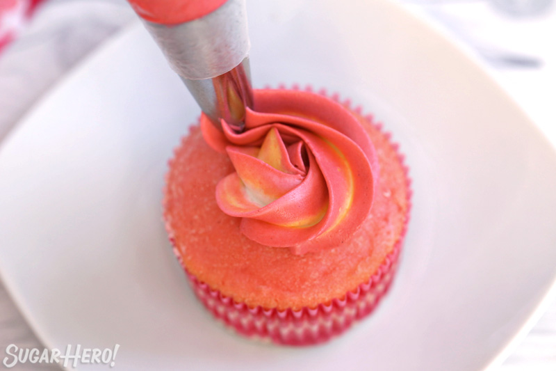 Beginning to pipe a pink and yellow swirl on top of a pink cupcake to make a buttercream rosette.