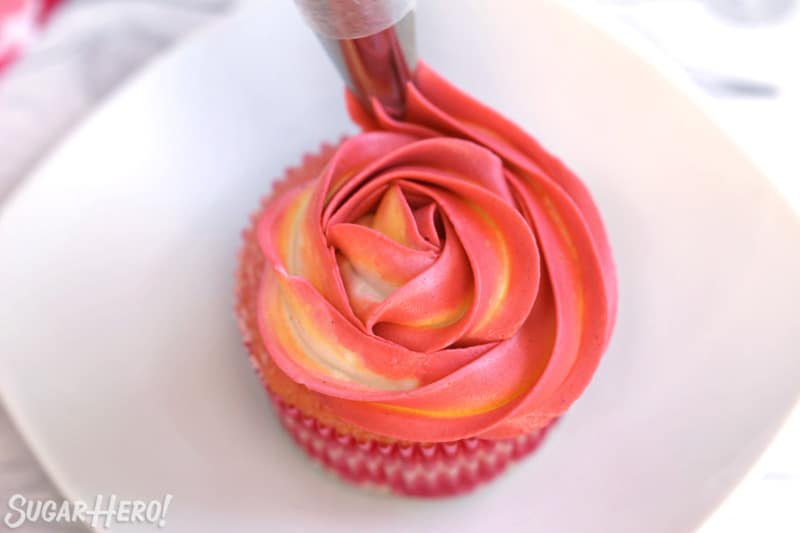 Finishing piping the swirl on a pink-yellow rosette atop a cupcake.