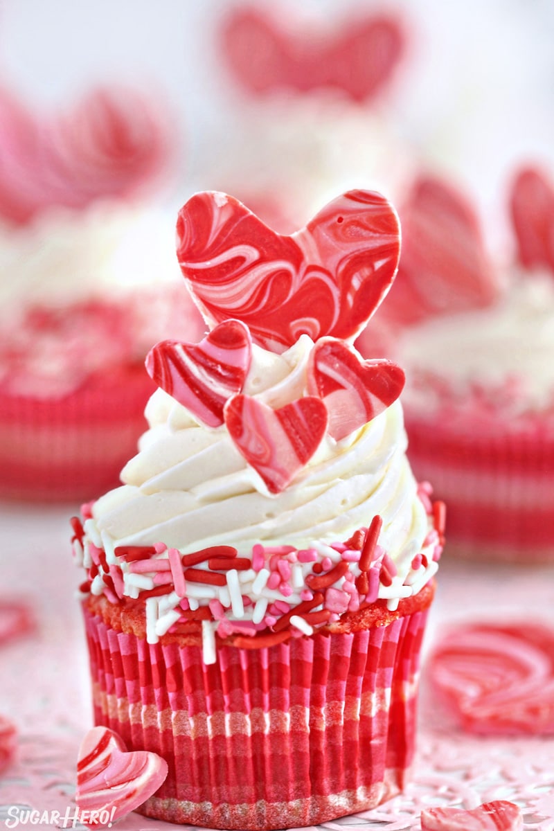 Close-up of cupcake with white buttercream, pink and red sprinkles, and four chocolate hearts on top.