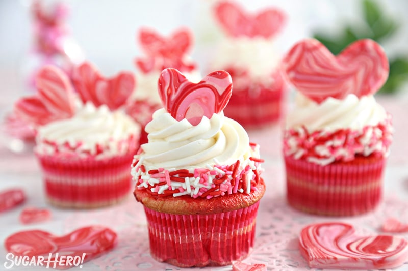 Group of red velvet cupcakes with white buttercream and pink and red hearts on top.