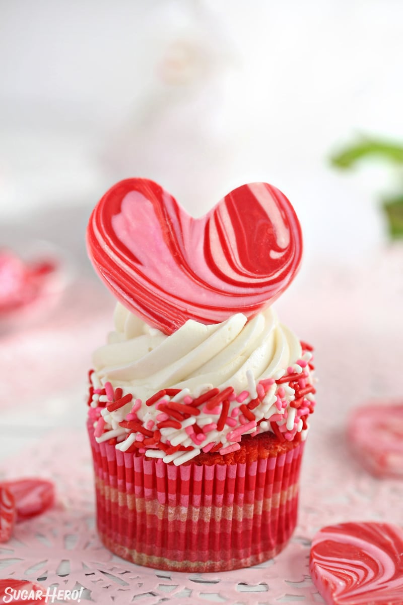 Close-up of a red velvet cupcake with white buttercream, sprinkles around the edge, and a chocolate heart on top.