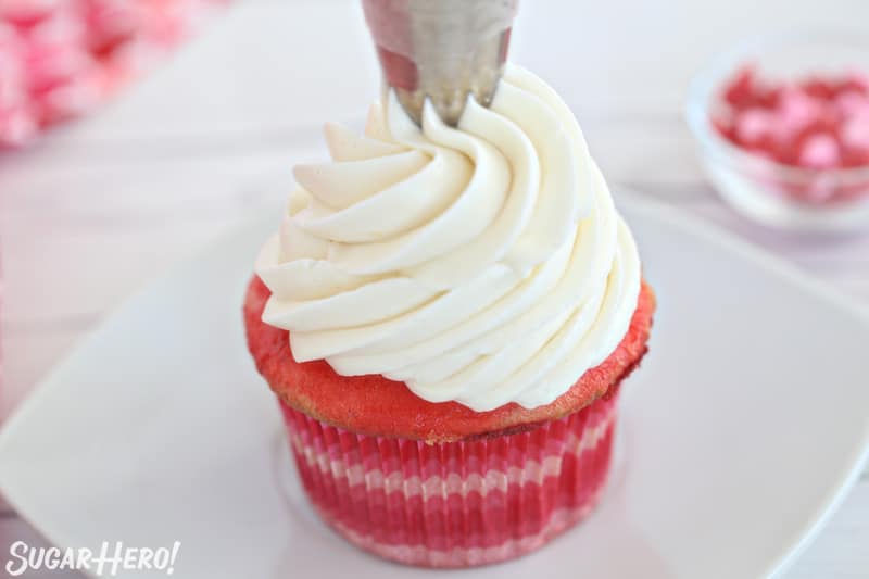 Piping a tall swirl of white buttercream on top of a pink cupcake.