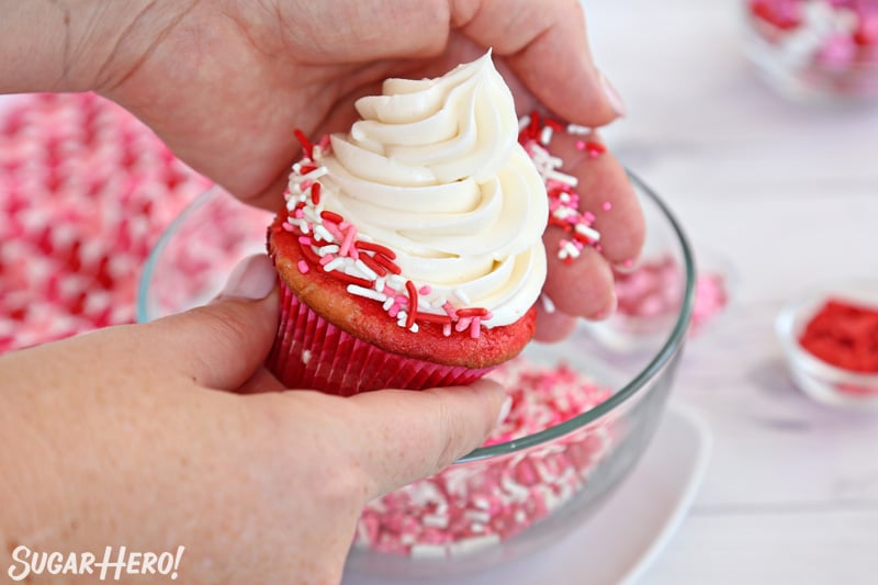 Hand pressing red, white, and pink sprinkles around the edge of a buttercream-frosted cupcake.