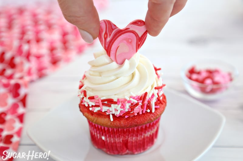 Hand placing a swirled chocolate heart on top of a buttercream-swirled cupcake.