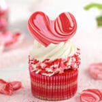 Close-up of cupcake with white buttercream, pink and red sprinkles, and a large chocolate heart on top.