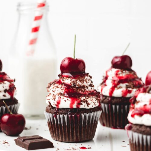 Several Black Forest Cupcakes on a white surface next to cherries and chocolate.