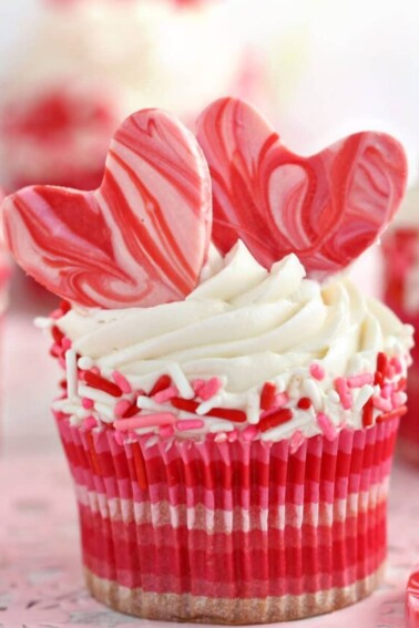 Close-up of cupcake with white buttercream, pink and red sprinkles, and two chocolate hearts on top.