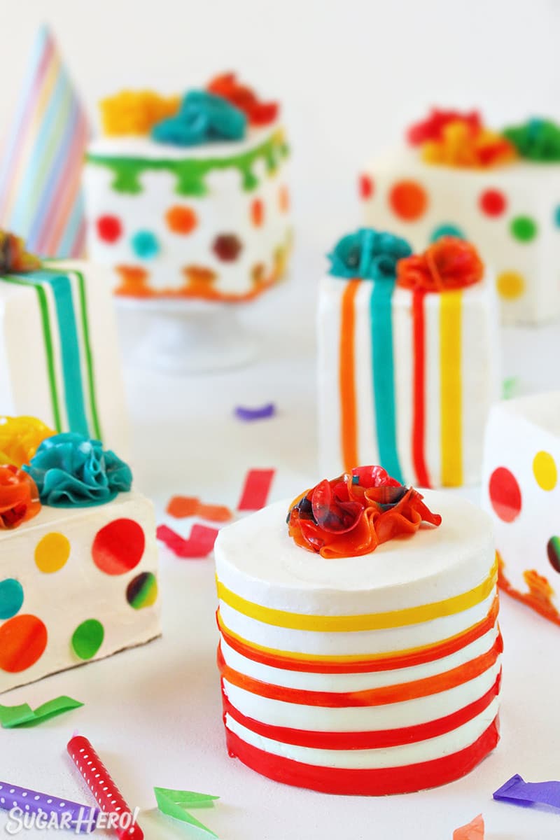 Group of multi-colored Birthday Present Mini Cakes on white background with confetti and candles scattered around.