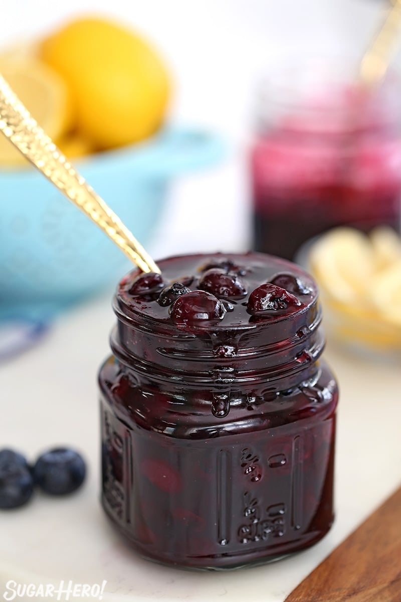 A glass jar full of blueberry sauce with a gold spoon in it.