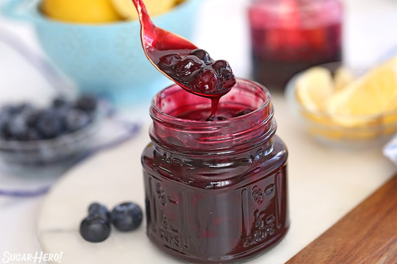 A glass jar of blueberry sauce with a spoonful hovering over the jar, various ingredients in the background