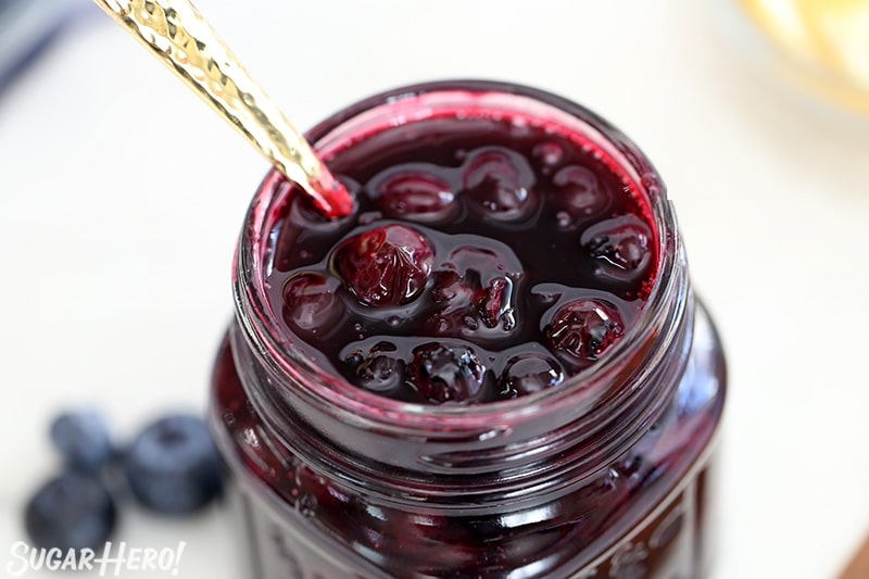 Close up of blueberry sauce in a glass jar with a gold spoon in the jar.