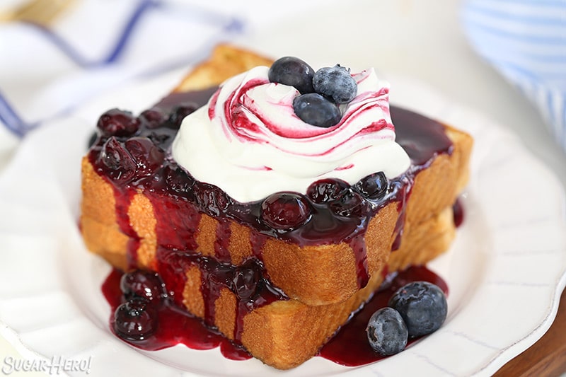 French toast covered in blueberry sauce and topped with whipped cream on a round white plate.