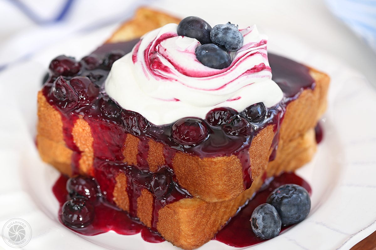 French toast covered in blueberry sauce and topped with whipped cream on a round white plate.