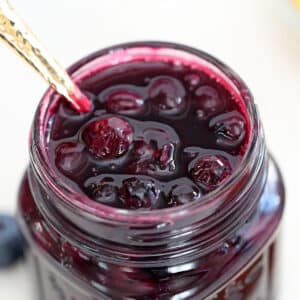 Close up of blueberry sauce in a glass mason jar with a gold spoon in it.