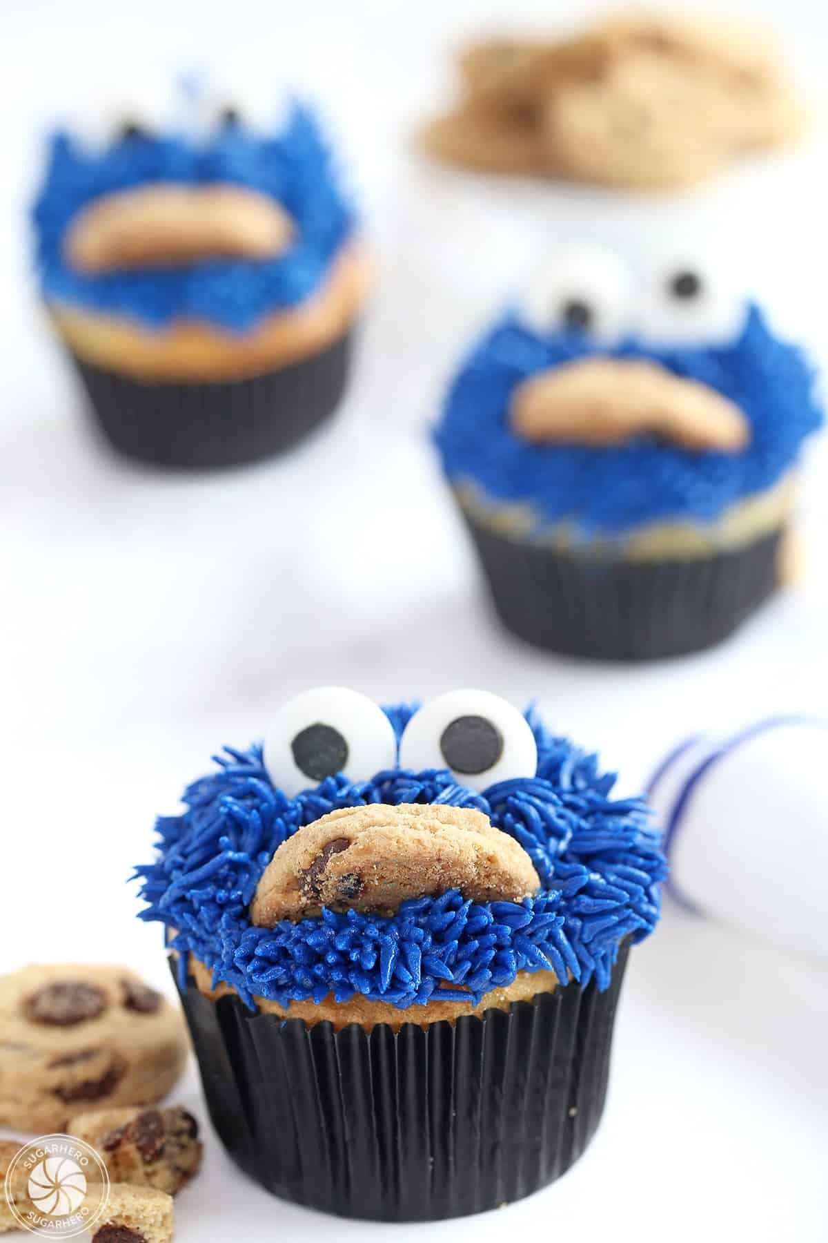 Three Cookie Monster Cupcakes on a white surface with crumbled cookies scattered around it.