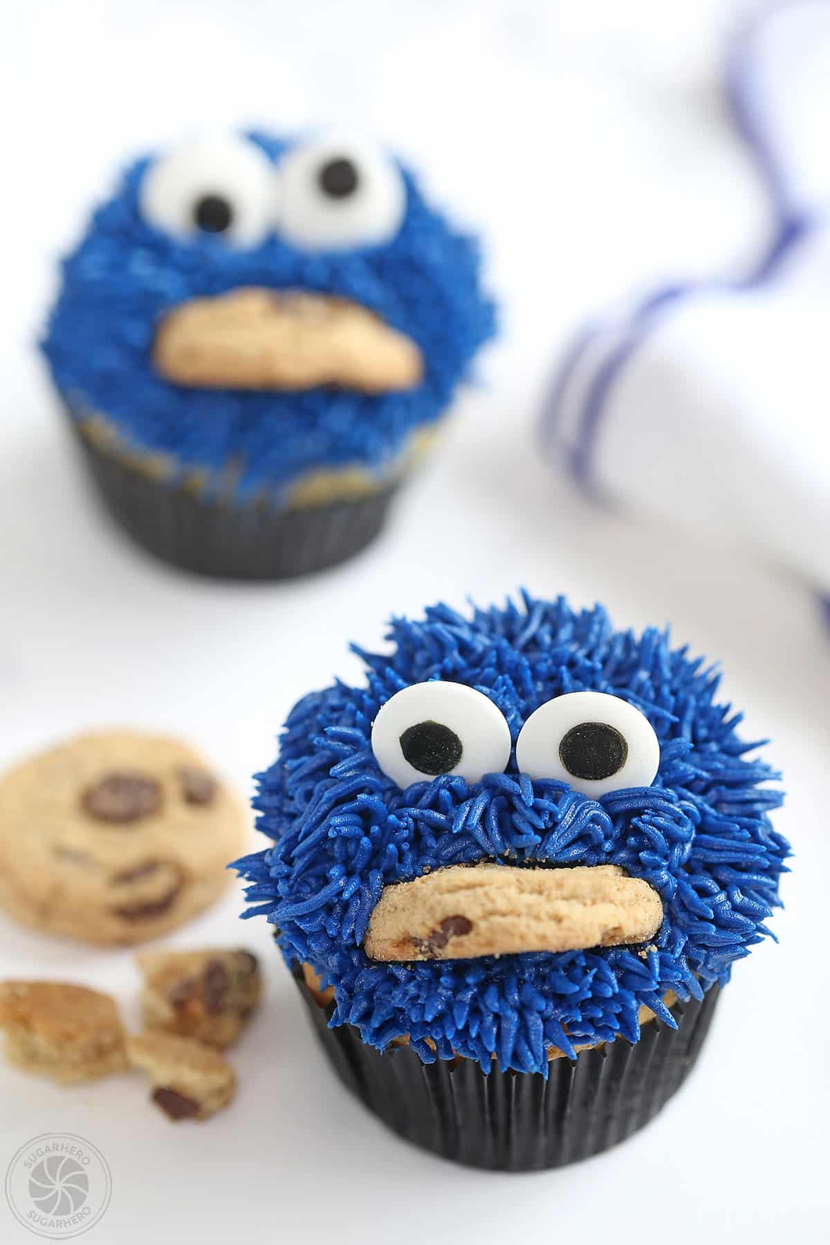 Two Cookie Monster Cupcakes with a white and blue napkin and crumbled cookies on the side.