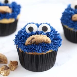 Three Cookie Monster Cupcakes on white surface with crumbled cookies on the side.