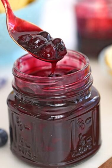 A glass jar of blueberry sauce with a spoonful hovering over the jar, various ingredients in the background