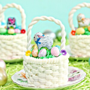 Five Easter Basket Cupcakes, each on small round plates, sitting on a bed of grass with Easter eggs scattered around.