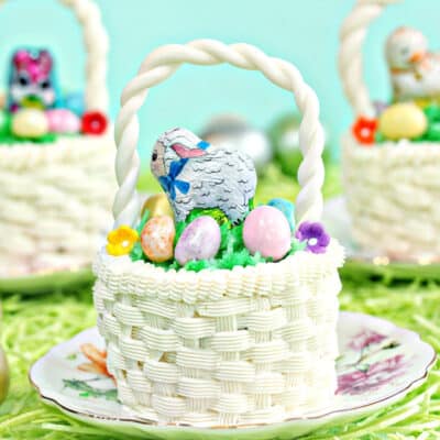 Three Easter Basket Cupcakes, each on small round plates, sitting on a bed of grass with Easter eggs scattered around.