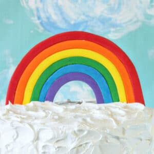 Fondant Rainbow Cake Topper on white frosting in front of a blue cloud background.