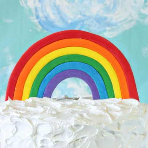 Fondant Rainbow Cake Topper on white frosting in front of a blue cloud background.