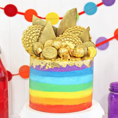 Gold-Topped Rainbow Cake on a white cake stand in front of colorful bunting in the background.