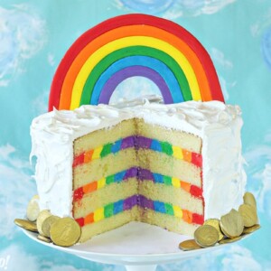 Cake cut open to reveal rainbow frosting stripes inside, on a white cake stand with a blue cloud background.