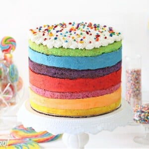 Rainbow Mousse Cake on a white cake stand with colorful lollipops and sprinkles around it.