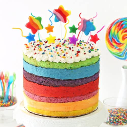 Rainbow Mousse Cake on a white cake stand with colorful lollipops and sprinkles around it.