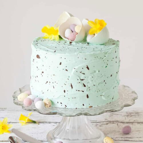 Layer cake with speckled light green frosting and Easter egg candies, egg shells, and yellow flowers on top.