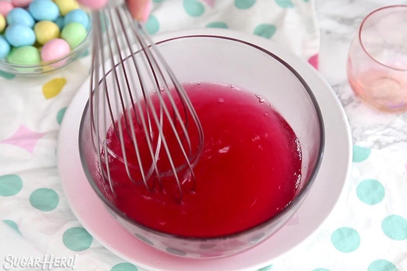 Whisking boiling water with red gelatin in a glass bowl.